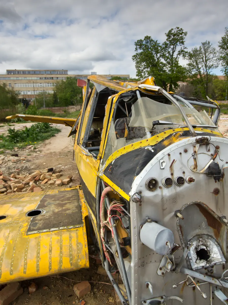 A severely damaged yellow aircraft fuselage, missing its engine, sits on a dirt lot next to scattered rubble. Wires hang exposed from the nose cone.