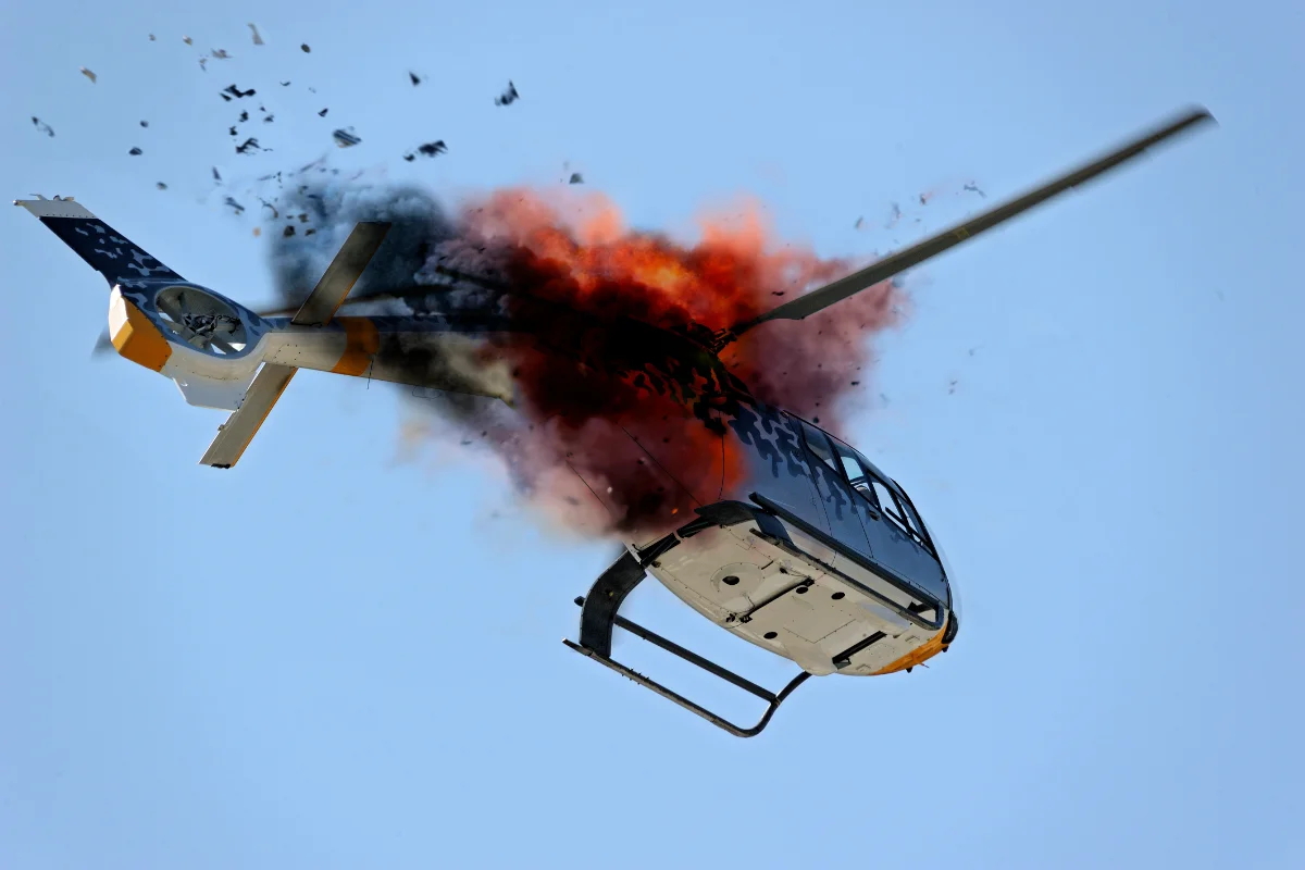 A helicopter is exploding mid-flight against a blue sky, with bright orange flames and thick black smoke billowing from its fuselage.
