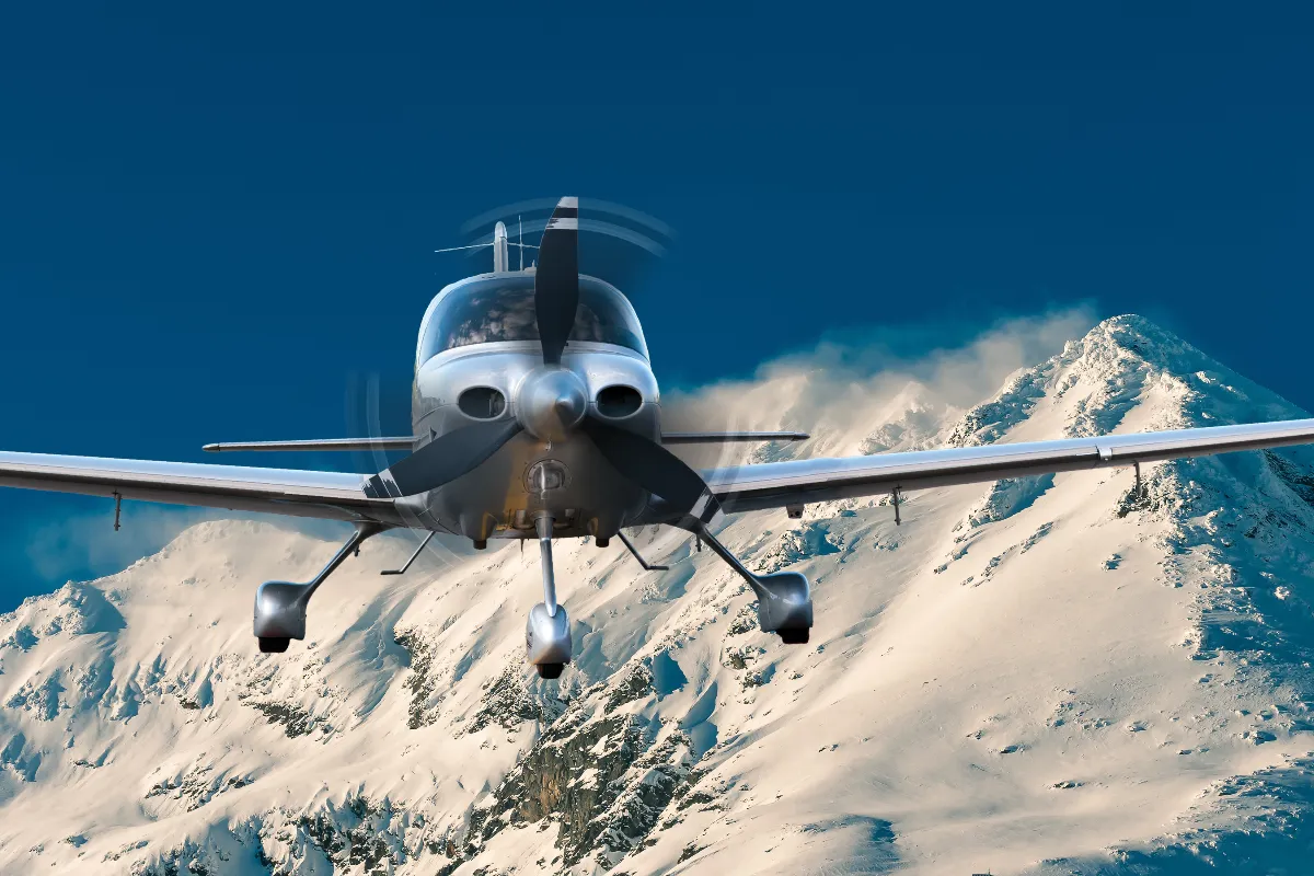 A small, single-engine silver airplane flies directly toward the camera, its propeller spinning, above a vast expanse of snowy mountains under a deep blue sky.